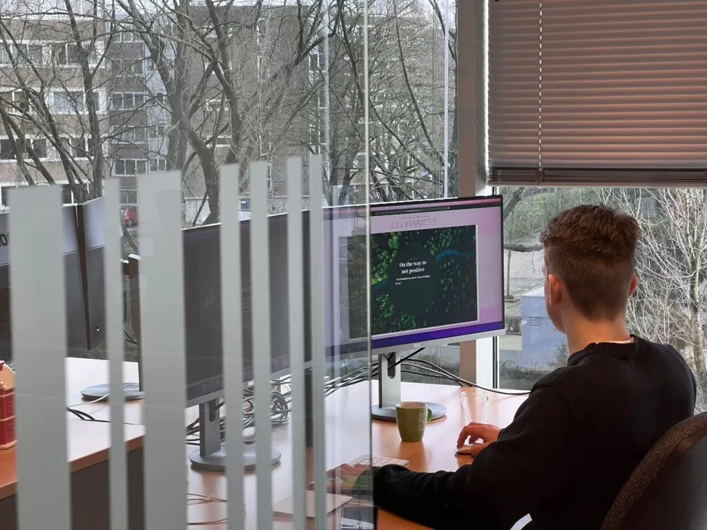 A developer at a monitor behind a glass partition, bare trees through the window beyond.
