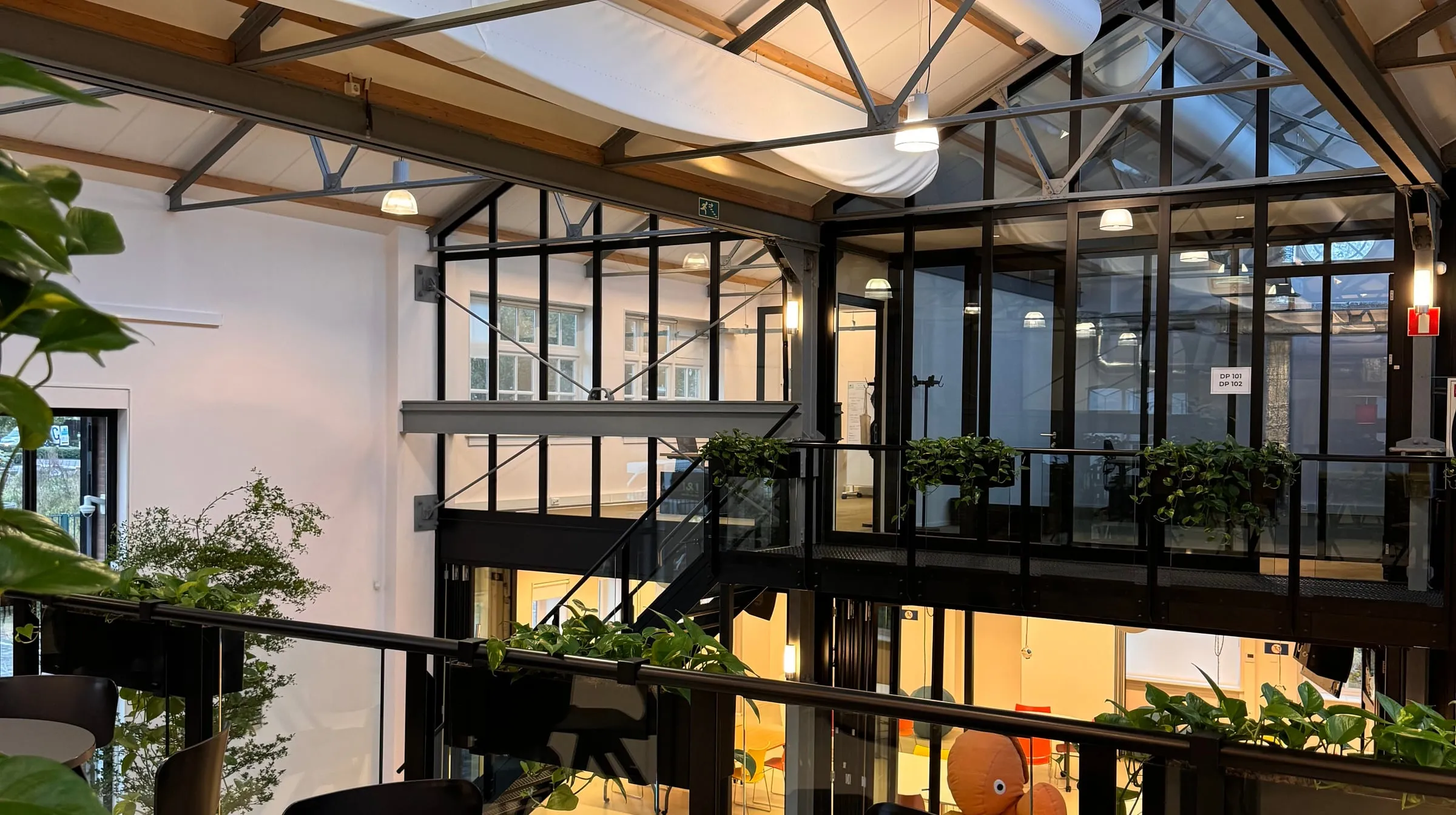 The Consent Studio office in Tilburg: a converted warehouse with a steel and glass mezzanine, plants along the rail, and tables on the floor below.