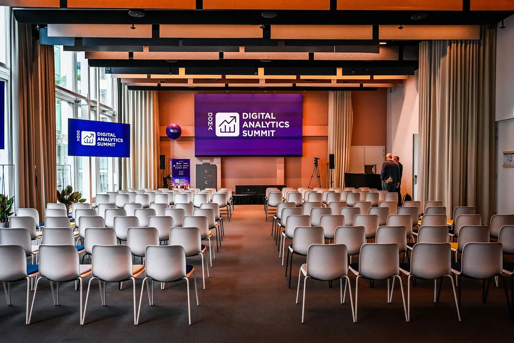 Rows of empty white chairs facing a stage, the Digital Analytics Summit logo on the screen behind it.